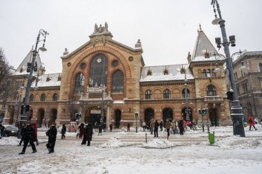 Macaristan 'ın Budapeşte kentindeki tarihi Büyük Pazar Salonu' nu şiddetli bir kar fırtınasının ortasında gösteren büyüleyici kentsel kış sahnesi. Görüntü, karla kaplı sokaklar, küreklenmiş beyaz kar ve yayalar ile şehrin atmosferik soğuğunu yakalıyor.