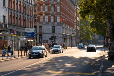Slovenya, Ljubljana 'nın canlı kent nabzını yakalayan bu görüntü, tarihi Grand Hotel Union yakınlarındaki işlek bir sokak sahnesini gösteriyor. Yayalar kaldırımda yürürken, araçlar asfalt yolda otobüs şeridi ile işaretlenmiş şekilde ilerliyorlar. Sıcak altın saat güneşi