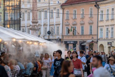 Mestni Trg 'de turistlerin ve yerlilerin restoran terasında su altında serinlediği canlı bir şehir manzarası. Arkaplanda tarihi Robba Çeşmesi ve barok mimarisi yer alıyor. Yaz seyahatleri ve sıcak hava dalgaları için mükemmel.