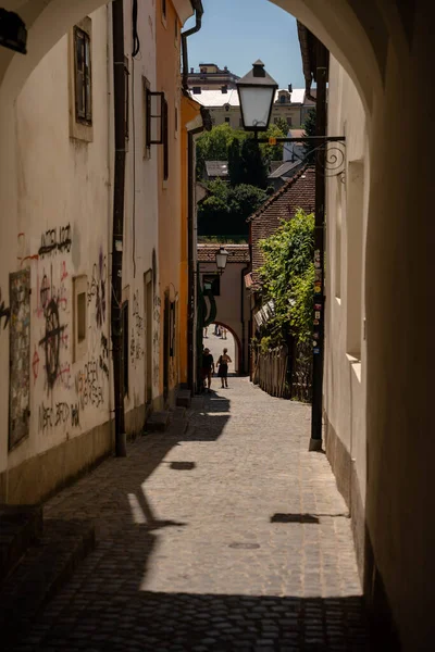 Slovenya 'nın Maribor kentindeki güneşli bir kaldırım taşına gölgeli bir kemerden bakan pitoresk dikey bir manzara. Dar geçitte yıpranmış duvarlar, kentsel grafiti, eski bir demir sokak lambası ve sürünen sarmaşıklar yer alıyor. İki yaya yürüyor.