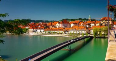 Maribor, Slovenya - 9 Ağustos 2025: Maribor 'un Drava Nehri boyunca uzanan Paskalya Köprüsü' nü ve turuncu çatılı evleri canlı mavi gökyüzü altında gösteren parlak bir zaman çizelgesi ele geçirdi.