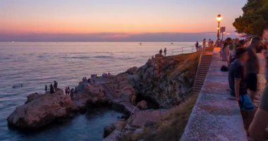 Rovinj 'deki akşam sahnesinde insanlar sıcak sokak ışıklarıyla sahil kenarındaki gezinti alanında pastel gün batımını izlemek için toplanıyor..