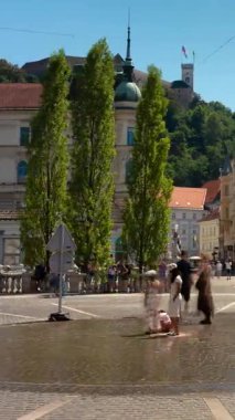 Ljubljana, Slovenya - 10 Ağustos 2025: Zaman ayarlı, kaydırmalı geçiş, dikey. Slovenya 'daki Triple Bridge ve Ljubljana Castle View' de Çocuklar Yer Pınarında Oynuyor ve Preseren Meydanı 'nda Bulanık İnsanlar Canlı Bir Yaz Sahnesi