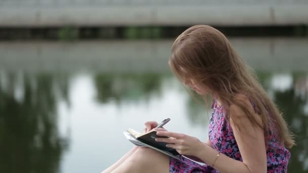 La hermosa joven escribe algo en su cuaderno en la orilla del río — Vídeos de Stock