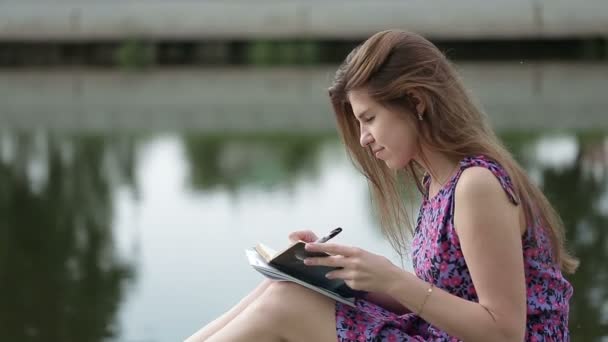 Tânăra femeie scrie ceva în caietul ei de pe malul râului — Videoclip de stoc