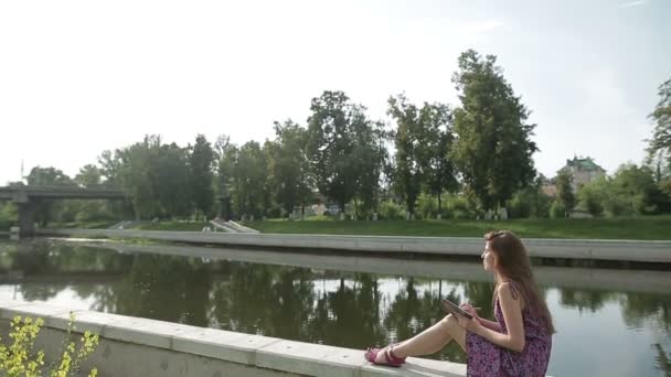 Mujer joven escribe algo en su cuaderno en la orilla del río en el parque de la ciudad — Vídeos de Stock