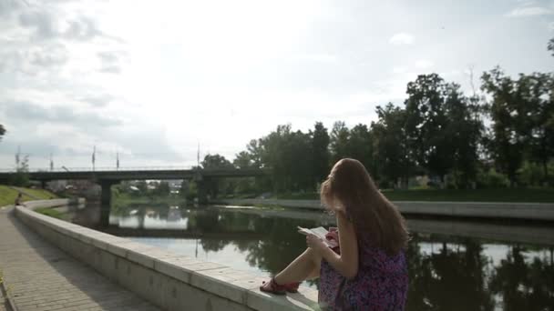 Bella donna scrivere qualcosa nel suo quaderno sulla riva del fiume nel parco della città — Video Stock