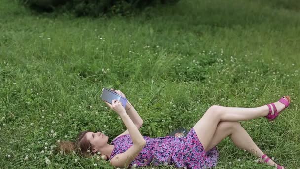 Chica joven leyendo un libro en el parque mientras está acostado en la hierba — Vídeos de Stock