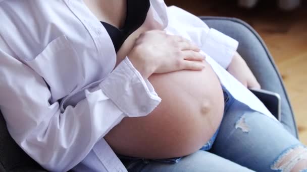 Νεαρή έγκυος γυναίκα χαϊδεύει μεγάλη κοιλιά στην πολυθρόνα. — Αρχείο Βίντεο