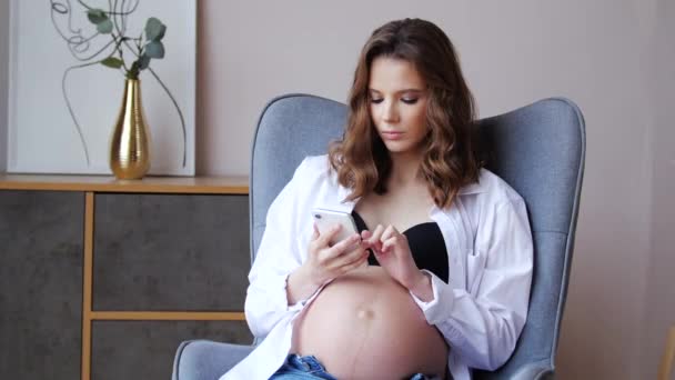 Retrato de mujer embarazada de belleza con el pelo largo usando el teléfono en el sillón en casa — Vídeos de Stock