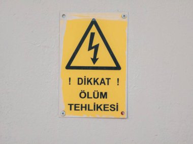 Gri bir duvarda sarı ve siyah elektrikli tehlike uyarısı var. İşarette bir yıldırım sembolü ve Dikkat Olum Tehesi adlı Türk metni yer alıyor, bu da yüksek voltajı işaret eden Uyarı, Ölüm Tehlikesi anlamına geliyor.