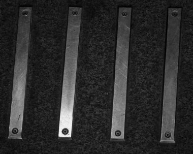 A minimalist black and white photograph showing a close-up of four parallel metal strips. The scratched metal bars are screwed into a dark, rough, textured surface, creating a strong abstract pattern with an industrial aesthetic.