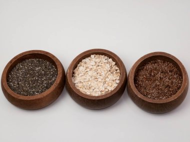 Three dark wooden bowls filled with healthy breakfast ingredients. The bowls contain chia seeds, rolled oats, and flax seeds, arranged in a row on a clean white background. A concept for superfoods, healthy eating, and balanced nutrition.