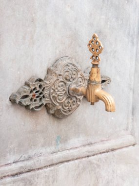 Yıkanmış mermer bir duvara monte edilmiş, karmaşık detayları olan dekoratif antika bir pirinç musluğun yakın çekimi. Bu geleneksel halka açık çeşme musluğu tarih, kültür ve eski dünya mimarisini çağrıştırıyor..