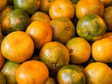 Büyük bir taze mandalina yığınının yakın çekim görüntüsü. Meyveler canlı turuncu ve yeşil renklerin karışımını sergiliyor, olgunluğun çeşitli aşamalarını gösteriyor ve dokulu bir arkaplan oluşturuyor..