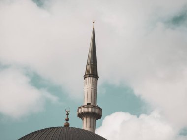 Görkemli bir caminin minare ve kubbesinin alçak açılı görüntüsü. Geleneksel İslami mimaride, dingin, bulutlu bir gökyüzünün zeminine kurulmuş uzun bir kule ve bir altın hilal sembolü yer alıyor ve bu da barış ve ruhanilik hissi uyandırıyor..