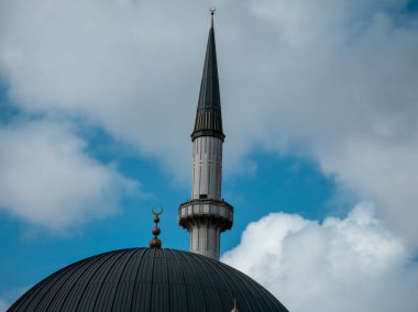 Bulutlu bir gökyüzüne karşı bir cami kubbesi ve minarenin yakın görüntüsü. Mimari detaylar arasında, her ikisi de İslami inanç ve kültürü temsil eden hilal şeklinde altın sembollerle süslenmiş tırtıklı bir kubbe ve uzun minare yer alıyor..