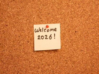 'Hoş geldiniz 2026!' diye el yazısıyla yazılmış bir mesaj. Sarı yapışkan bir notta. Not kahverengi desenli bir mantar panoya kırmızı bir iple tutturulmuş. Yeni Yıl, gelecek, planlama ve 2026 yılı beklentisi.