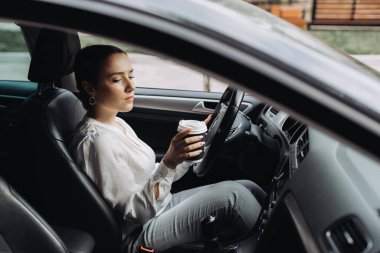 Kadın kahve içiyor ve arabada telefonunu kontrol ediyor. Trafik sıkışıklığında duruyor.