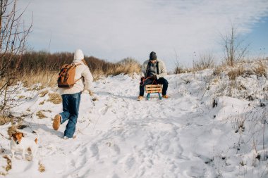 Baba kızlarının ellerini, karlı bir tepede nefesiyle ısıtıyor. Açık havada, iç açıcı bir aile anının tadını çıkarıyor.