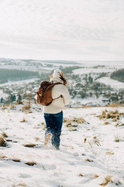 Sırt çantalı ve küçük köpekli bir insan, karlı arazide kış yürüyüşünün tadını çıkarıyor, kırsal bir tepeden aşağı iniyor.