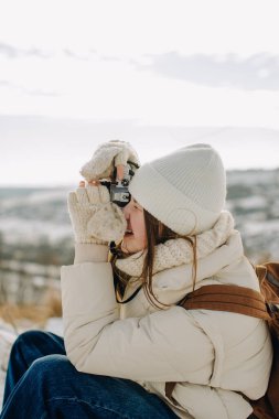 Sıcak giysiler içinde oturan, açık hava kış macerasının tadını çıkaran ve klasik bir kamerayla resim çeken genç bir kadın.