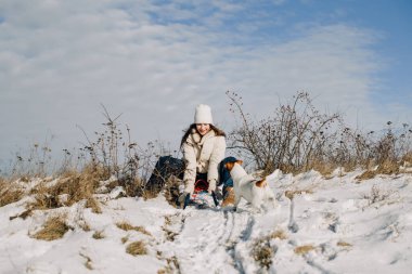 Genç bir kadın kışın bir kızağın üzerinde otururken gülümsüyor, açık havada küçük bir köpekle oynuyor.