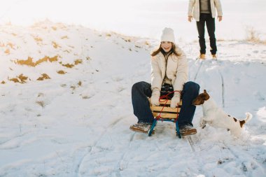 Kız gülümsüyor, tahta bir kızağın üzerinde oturuyor ve yanında köpeğiyle karda zıplıyor. Babam arka planda duruyor.