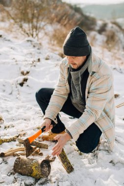 Man splitting logs with an axe on snowy ground, preparing wood for a winter campfire in the wilderness