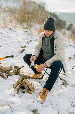 Kışın giyinmiş bir adam, ıssız bir yerde gaz lambası kullanarak kamp ateşi yakmaya hazırlanıyor.