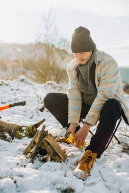 Sıcak giysiler içinde, soğuk, karlı kış manzarasında şömineyi gaz ocağı ve yakacak odunla yakmaya hazırlanan bir adam.