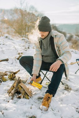 Karlı kış doğasında ısınmak için küçük kamp ateşini gaz lambasıyla yakan adam. Soğuk havada dışarıda kamp yapmanın keyfini çıkarıyorum.