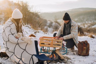 Karlı arazide kamp ateşiyle aile bağları, baba yemek hazırlarken kız battaniyeye sarılıyor.