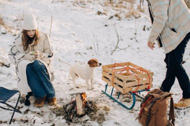 Aile kış gezisinde gevşiyor, kız çocukları köpekleriyle kamp ateşinde dinlenirken baba kızakla ayakta duruyor.