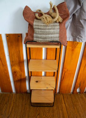 Siyah metal bir standın çerçevelediği ince ahşap bir rafın üzerinde, yumuşak giysili bir insan bacağının ipucuyla birlikte, desenli, toprak renginde bir dokuma torbası oturuyor..