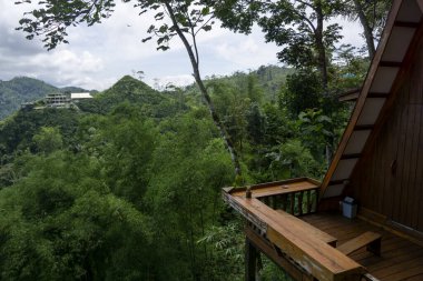 Bir dağ kulübesinin ahşap balkonu yoğun bir tropikal ormana doğru uzanır ve yeşil tepeleri ve uzak yapıları huzurlu bir şekilde görür. Seyahat, doğa ve geri çekilme temaları için ideal.