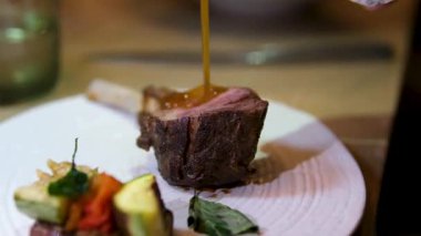 Altın karamel-kahverengi soslu orta pişmiş lama bifteğin yakın çekimi, canlı sebzelerle dolu beyaz bir tabakta servis edilir..