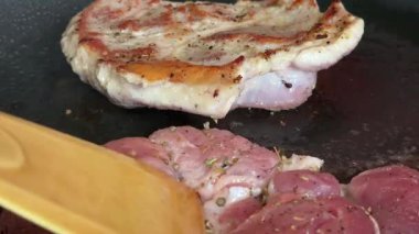 Ahşap bir spatula gibi siyah bir tavada kızaran iki domuz bifteği onları çevirir. Buharı serbest bırakır. Cızırdayan sulu eti ve iştah açıcı pişirme tarzını ön plana çıkarırlar..