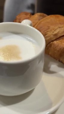 Beyaz fincanda kapuçino ve kruvasan var. Kadın eli masada duruyor ve kahveli fransız kahvesi içiyor. Yüksek kalite 4k görüntü