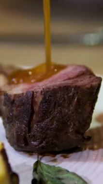 Altın karamel-kahverengi soslu orta pişmiş lama bifteğin yakın çekimi, canlı sebzelerle dolu beyaz bir tabakta servis edilir..