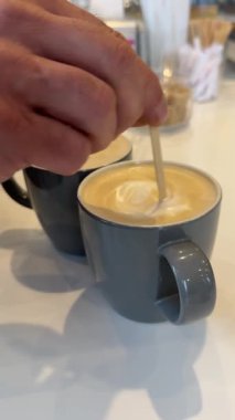 Erkek elleri, yumuşak bir kapuçinoyu metal bir kaşıkla yavaşça karıştırırken, bir kafede rahat bir anın tadını çıkarırken, rahat bir atmosferle çevrili sıcak içeceğin tadını çıkarıyor. Yüksek kalite 4k