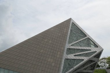 a large triangular building with a glass facade stands on the right side of an open square in front of it. the roof is made from gray ceramic tiles and has multiple angular patterns, giving it a futuristic feel