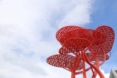 A red sculpture of a tree with a blue sky in the background. The tree is made of metal and has a lot of holes
