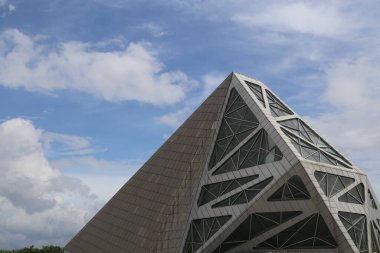a modern building in the shape of an angular pyramid, made from metal and glass with geometric patterns on its exterior walls. the sky is blue with white clouds, creating a contrast against the grey tile roof