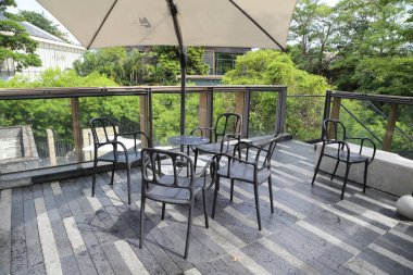 the outdoor terrace of the cafe features black steel chairs and tables, surrounded by greenery. the balcony is enclosed with glass railings offering views over the trees to distant buildings.