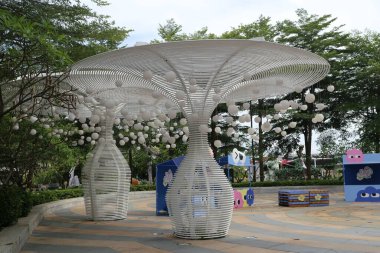 a white metal mesh structure with a mushroom-shaped roof and lanterns hanging from it is located in the middle of an outdoor park, surrounded by trees and children's play equipment, creating a colorful scene.