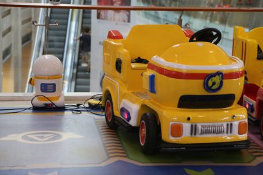 a children's cute yellow car toy, a machine with an electric motor, in the shape of a truck and a fire truck, is charging on the floor next to it inside a shopping mall. a small robot stands nearby for guidance.