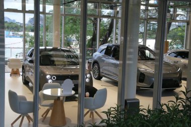 İçinde cam duvarlar ve içeriyi net görebilen geniş pencereler olan üç arabalı bir araba galerisi. Sahne, modern tasarım elementleriyle süslenmiş kahverengi bir aracın yanında iki gri renkli elektrikli araç içeriyor.