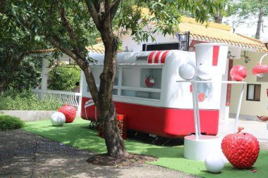 Beyaz ve kırmızı retro tarzı bir yemek kamyonu Amerikan tarzı bir villanın bahçesine park edilmiş, bir tarafında açık yeşil çimenler ve yanında çileğe benzeyen büyük kırmızı toplar var..