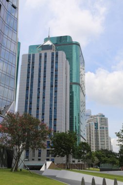 Sağında cam perdeli uzun bir bina, önünde yeşil çimenler ve ağaçlar, arkasında başka bir ofis kompleksi bulunur. Modern mimarisi ve yüksek binaları olan ticari bir cadde.